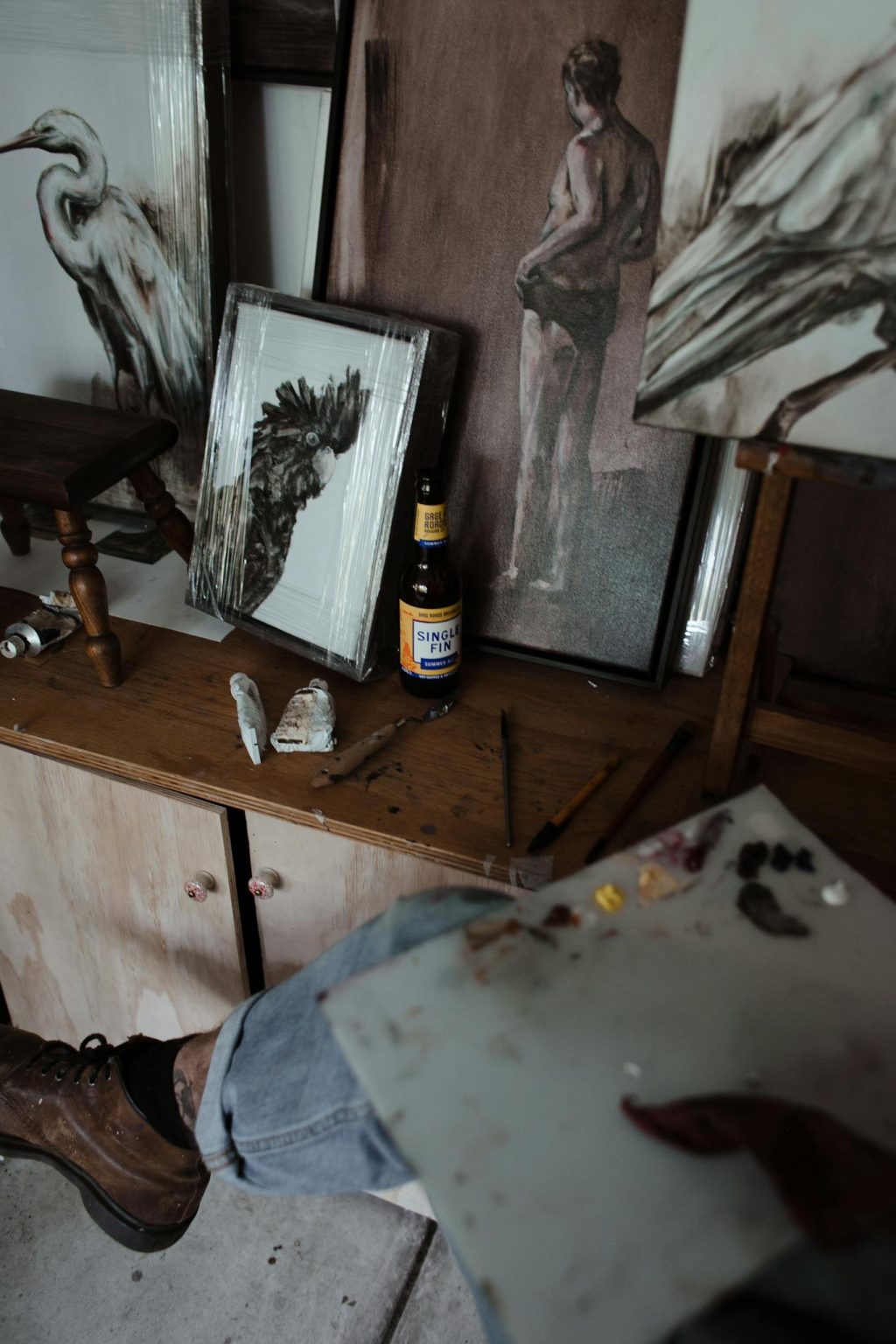 From above of crop anonymous male artist sitting on chair near wooden table with collection on various framed paintings in creative workshop