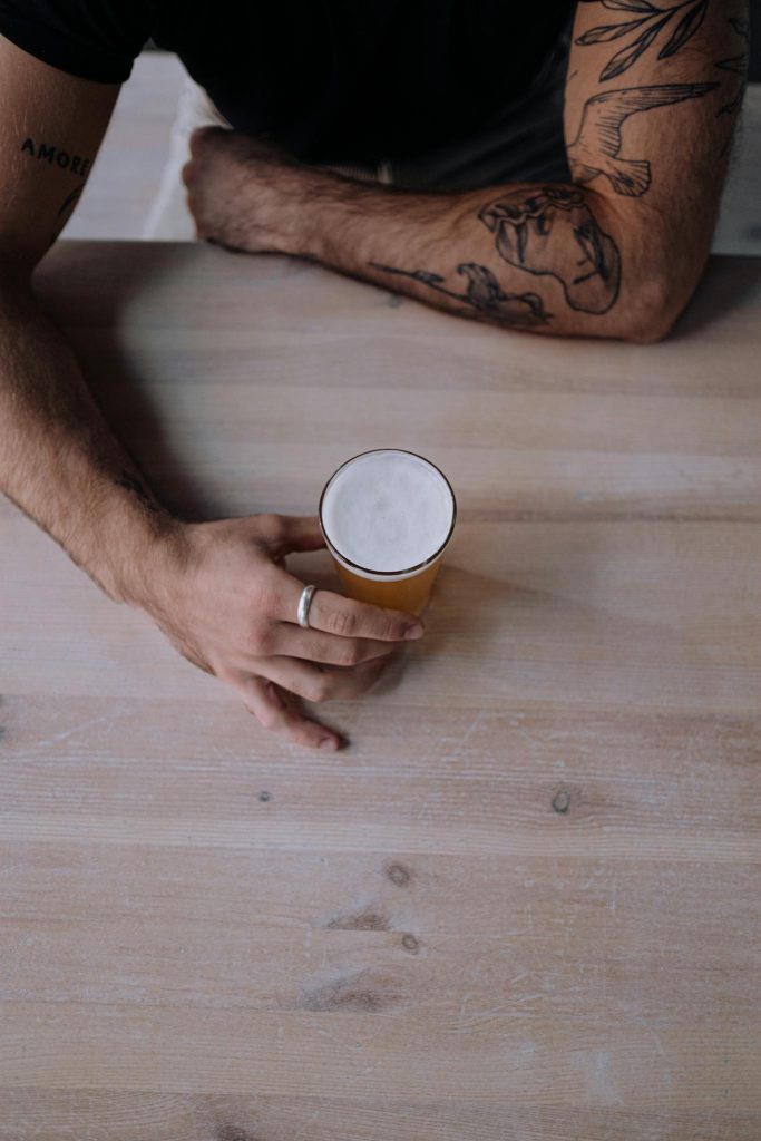 Overhead shot of a tattooed arm holding a craft beer on a rustic wooden table.